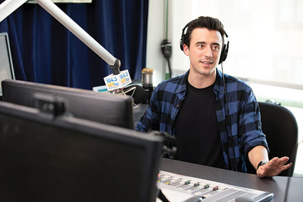 Jon Comouche sits in front of microphone and audio equipment